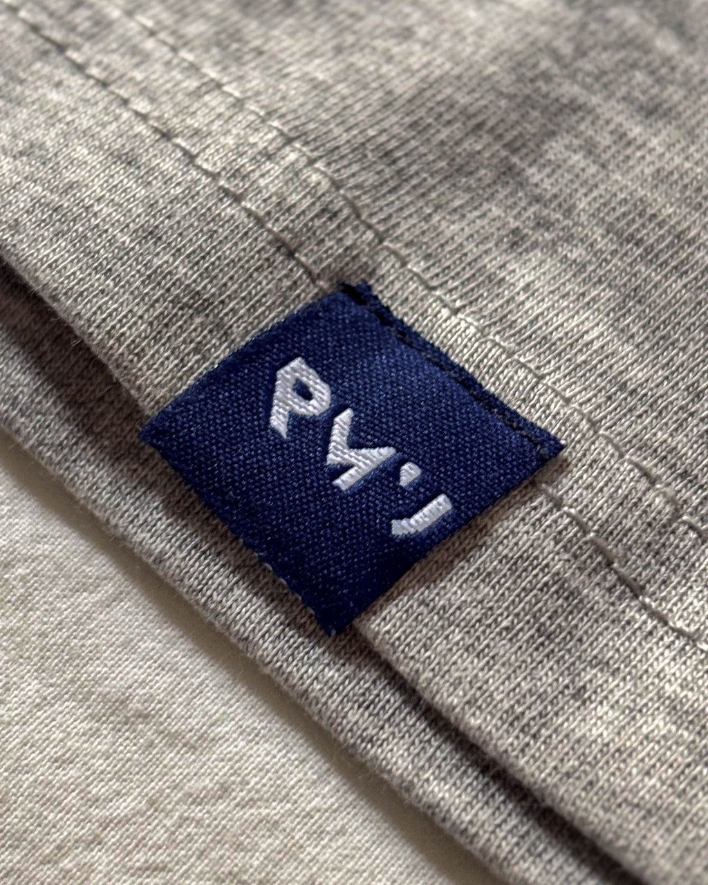 Detalle de la etiqueta azul navy RMU cosida en el bajo de la camiseta Core gris jaspeado
