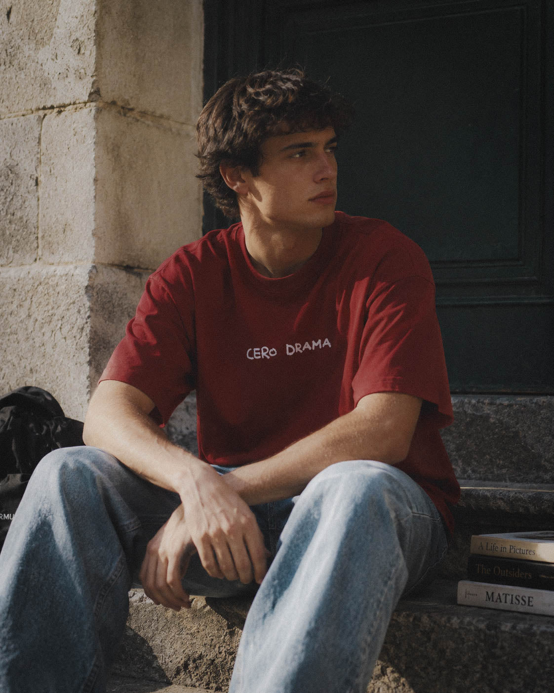 Chico con camiseta Cero Drama burdeos sentado en un portal